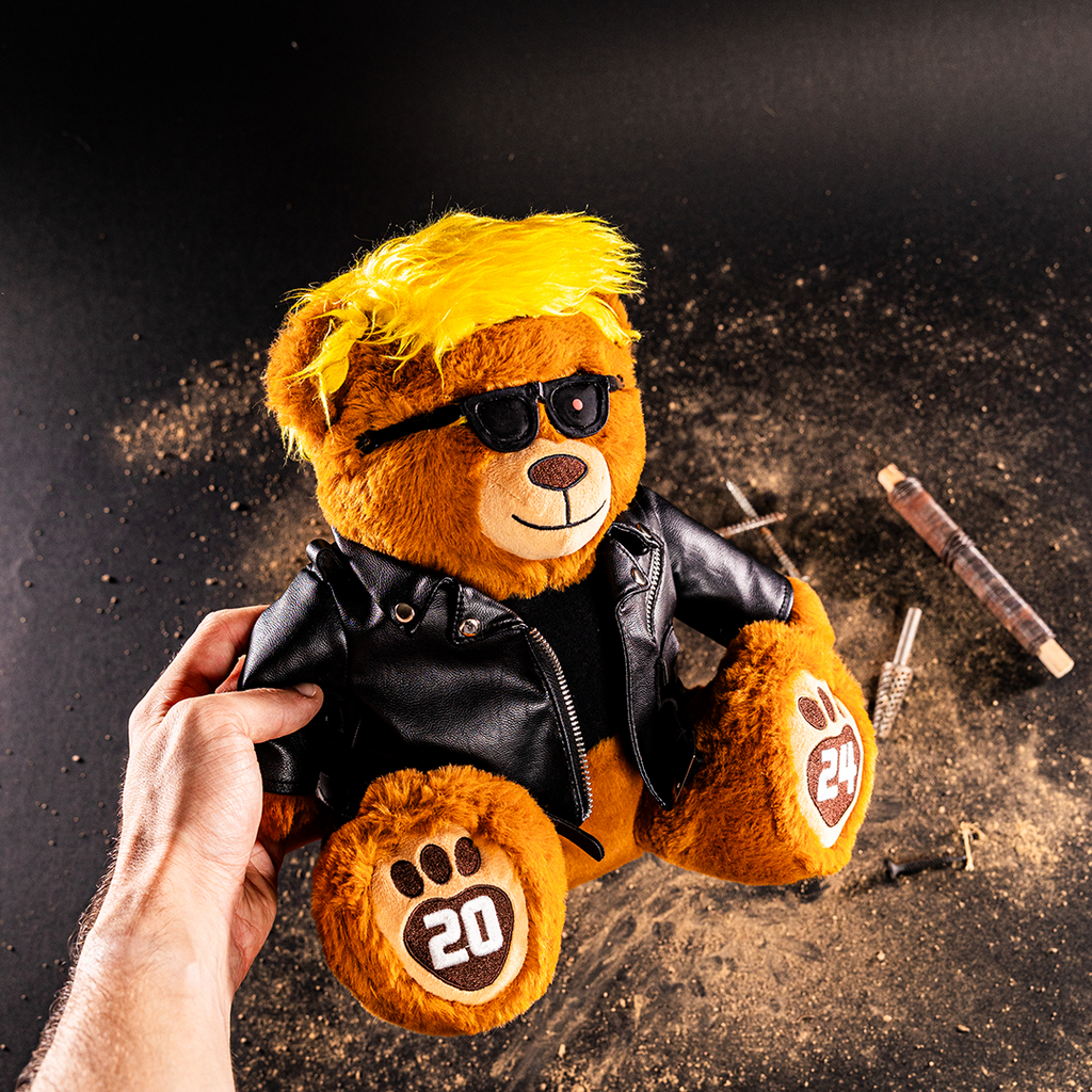 Trumpinator Teddy Bear with yellow hair, sunglasses, and a black leather jacket, holding a hand in a dusty background.