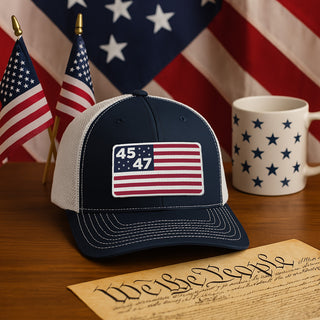 Navy blue trucker hat featuring a flag design with "45" and "47," placed on a wooden table with American flags and a mug.