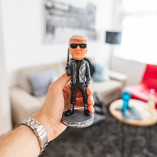 Trumpinator bobblehead held in hand, featuring sunglasses and a black leather jacket, set in a modern living room.