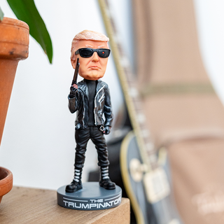 Trumpinator bobblehead in black leather outfit, holding a gun, with sunglasses, displayed on a wooden surface.