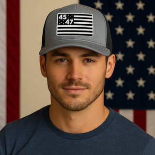 Gray monochrome hat featuring a 45th and 47th President flag design, worn by a man in front of an American flag backdrop.