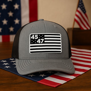 Gray and black trucker hat featuring a monochrome flag design with "45" and "47" on the front, placed on a wooden table.