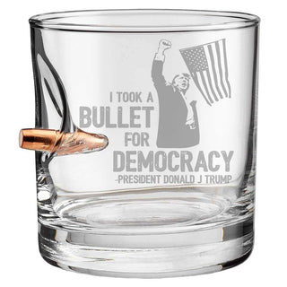 Trump whiskey glass featuring a bullet design and the phrase "I took a bullet for democracy" with a flag graphic.